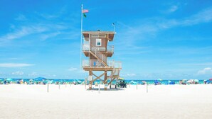 A lifeguard tower with a flag on top, located on a sandy beach with umbrellas and people in the background. - Unique Canal Home in The Best Location (Holmes Beach)
