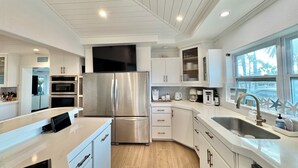 A modern kitchen with stainless steel appliances, white cabinetry, and a large window providing a view of palm trees. - Unique Canal Home in The Best Location (Holmes Beach)