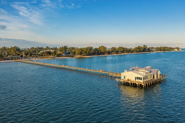 A waterfront property with a long pier extending into the water, a large house on stilts, and a beach in the background.