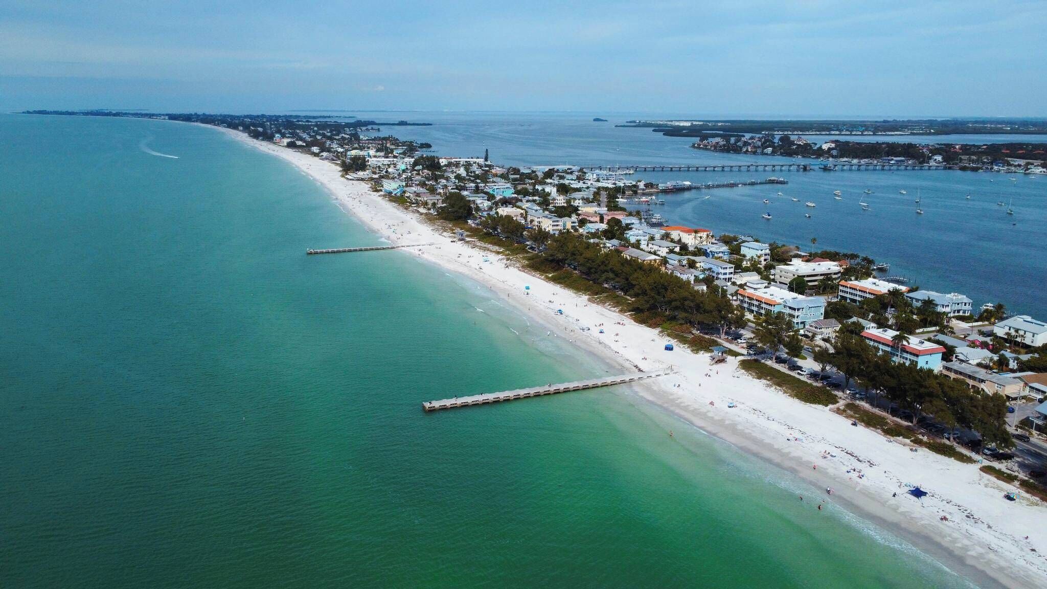 An aerial view of a coastal town with a long sandy beach, a pier, and buildings along the shore.