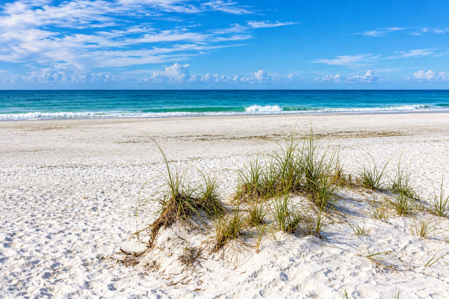 A beach with white sand and sparse grass, leading to the ocean under a clear sky.