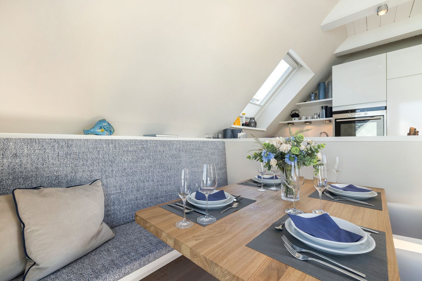 Ein Essbereich mit einem Holztisch, gedeckt für vier Personen, einem grauen Sofa und einer Küche im Hintergrund.