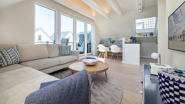 A modern living room with a sofa, coffee table, and chairs, a kitchen area with a dining table, and large windows offering a view of the cityscape. - Dünenloft - Relax and enjoy in the Dünenloft: your perfect vacation apartment on the (Rantum)