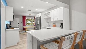 A modern kitchen with white cabinetry, a central island, and a dining area with wooden chairs. - Clearwater Pickleball,  Heated Pool, MiniGolf Oasis! 15 min to beach (Clearwater)
