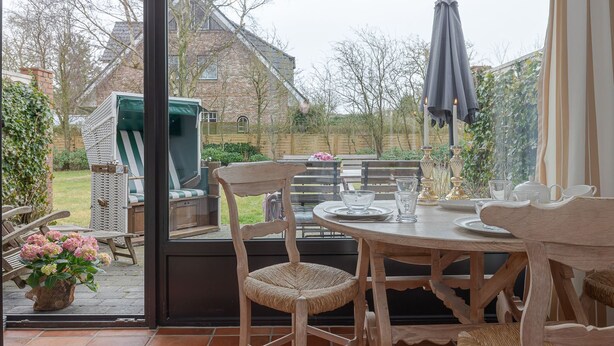 Une terrasse avec une table dressée pour les repas, des chaises en osier et un espace barbecue visible à travers la porte vitrée.