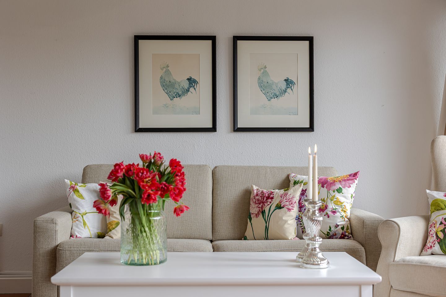 A living room with a beige sofa, floral cushions, a white coffee table, and two framed rooster artworks on the wall.