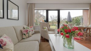 Un salon avec un canapé, une table ornée d’un vase de fleurs et, derrière une porte vitrée, une vue sur un espace patio extérieur.