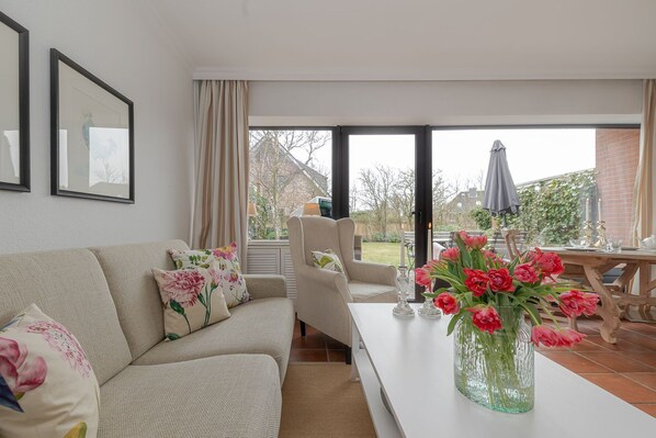 A living room with a sofa, a table with a vase of flowers, and a view through a glass door to an outdoor patio area.