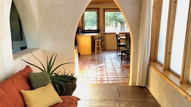 Um interior acolhedor com soalho de pedra, um sofá vermelho com almofadas, uma planta em vaso e vista para uma área de refeições através de um arco.