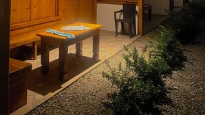 Un espace extérieur aménagé, abrité et doté de bancs et de tables en bois, entouré de verdure et recouvert d’un sol carrelé.
