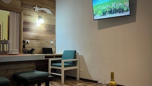 Une pièce moderne avec un mur en bois, un téléviseur diffusant des images du Costa Rica, un coin bar avec des tabourets, une table avec une bouteille et des verres, et un ordinateur portable posé sur une étagère.