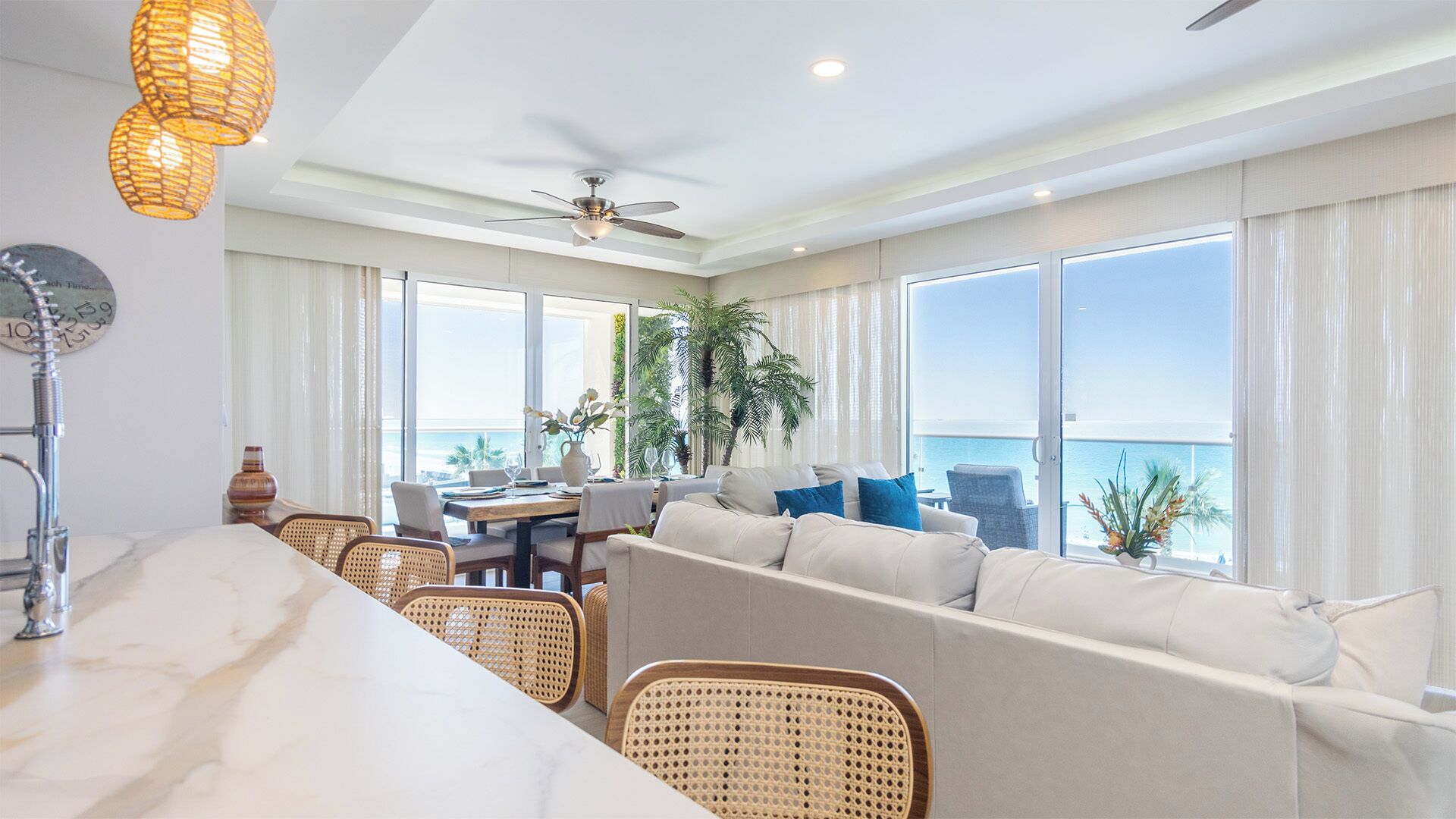 A spacious living area with a sofa, dining table, and chairs, large windows with a view of the ocean, a ceiling fan, and a decorative light fixture.