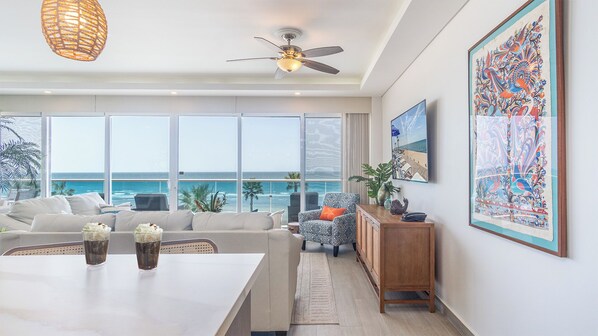 A modern living room with a large window overlooking the ocean, a ceiling fan, and a dining area with a table and chairs. - Unique condo by the beach at Encantame Towers C305 (Puerto Peñasco)