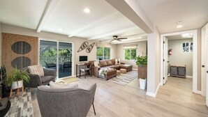 A spacious living area with a large sofa, a dining table, and a TV. There is a balcony with a view of greenery. - Lake View Home, 2 Spacious Decks, Fire Pit, WiFi, On Sight Casita Option Avail. (Lake Arrowhead)