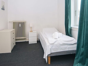 A hotel room with a bed, a bedside table with a lamp, a radiator, a dresser, and a window with curtains.