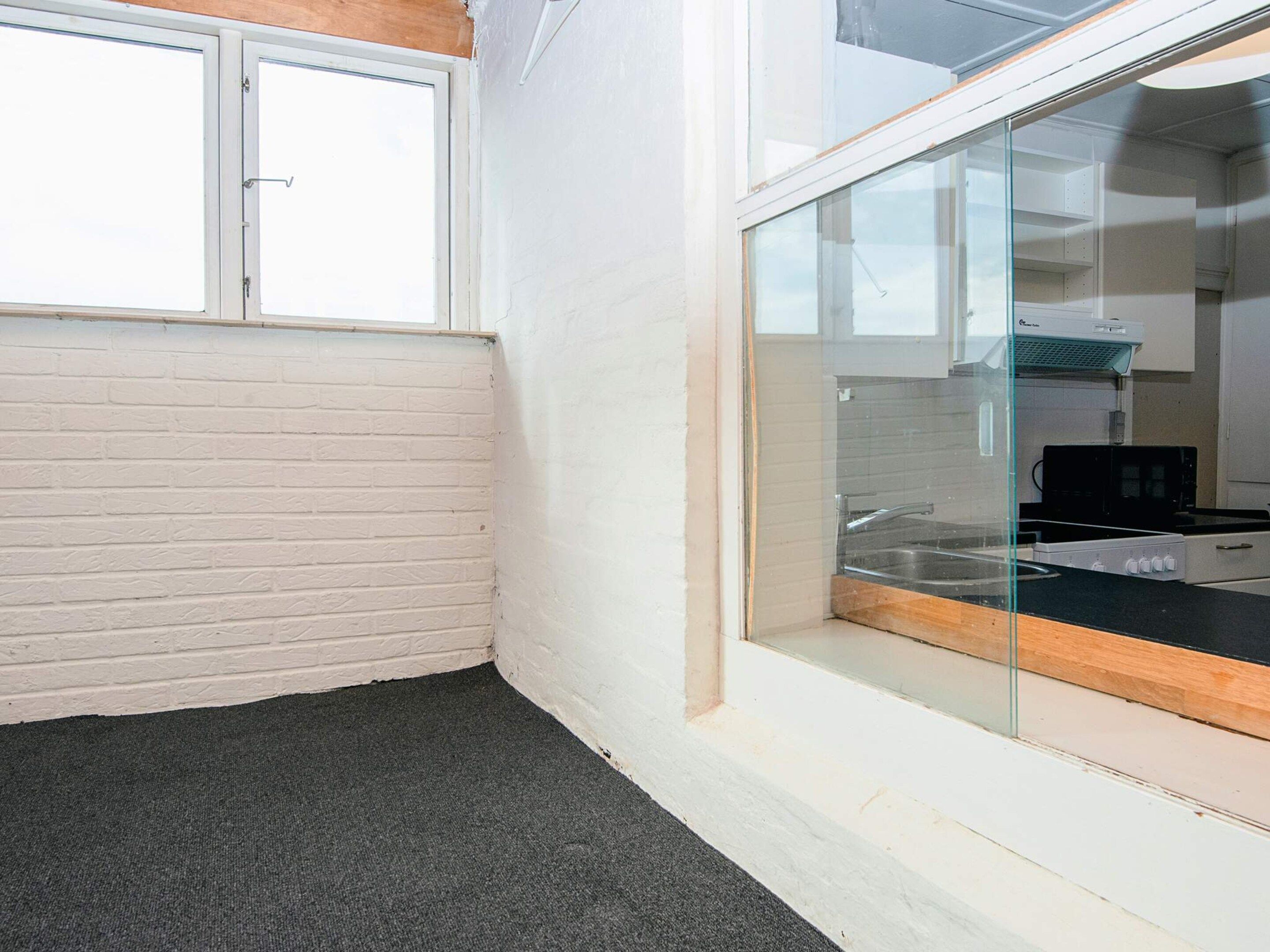 Un coin de pièce avec un mur en briques blanches, une moquette noire et une porte en verre donnant sur un espace cuisine équipé d’une cuisinière et d’un four à micro-ondes.