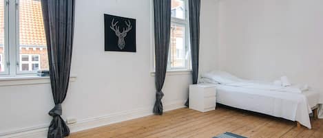 A bedroom with a bed, a bedside table, a nightstand, a window with curtains, and a wall-mounted picture.
