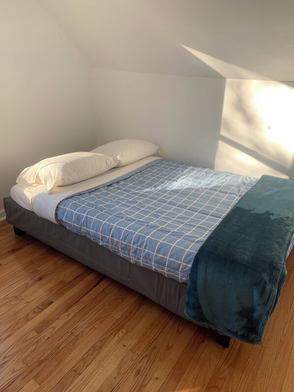 A single bed with a blue and white checkered bedspread, a teal throw blanket, and white pillows in a room with wooden flooring and a sloped ceiling.