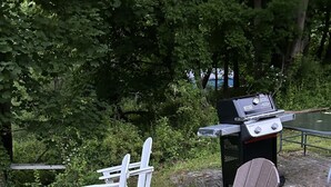 Montaje para picnic con sillas Adirondack y una parrilla en un césped con árboles al fondo.