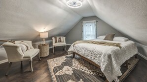 Un dormitorio acogedor en el ático, con techo inclinado, cama con colcha blanca, una silla, una mesita de noche con lámpara y una alfombra estampada.