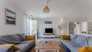 A modern living room with two grey sofas, a wooden coffee table, a flat-screen TV on a white stand, and a large window with a view of the outdoors. - Family home 2 steps from the Bassin (Gujan-Mestras)