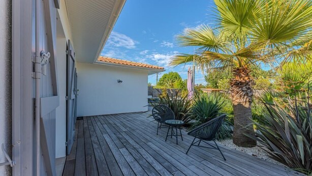 A wooden deck with outdoor furniture, a palm tree, and a building with a tiled roof. - Family home 2 steps from the Bassin (Gujan-Mestras)