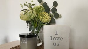 A candle labeled "The Woods Sand & Fog" on a woven mat, a small vase with green foliage, and a sign that says "I love us." - Off grid tiny house bush escape (Nar Nar Goon North)