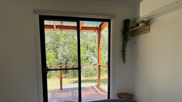 A bedroom with a bed, a sliding glass door leading to an outdoor deck, and a small wooden stool. - Off grid tiny house bush escape (Nar Nar Goon North)