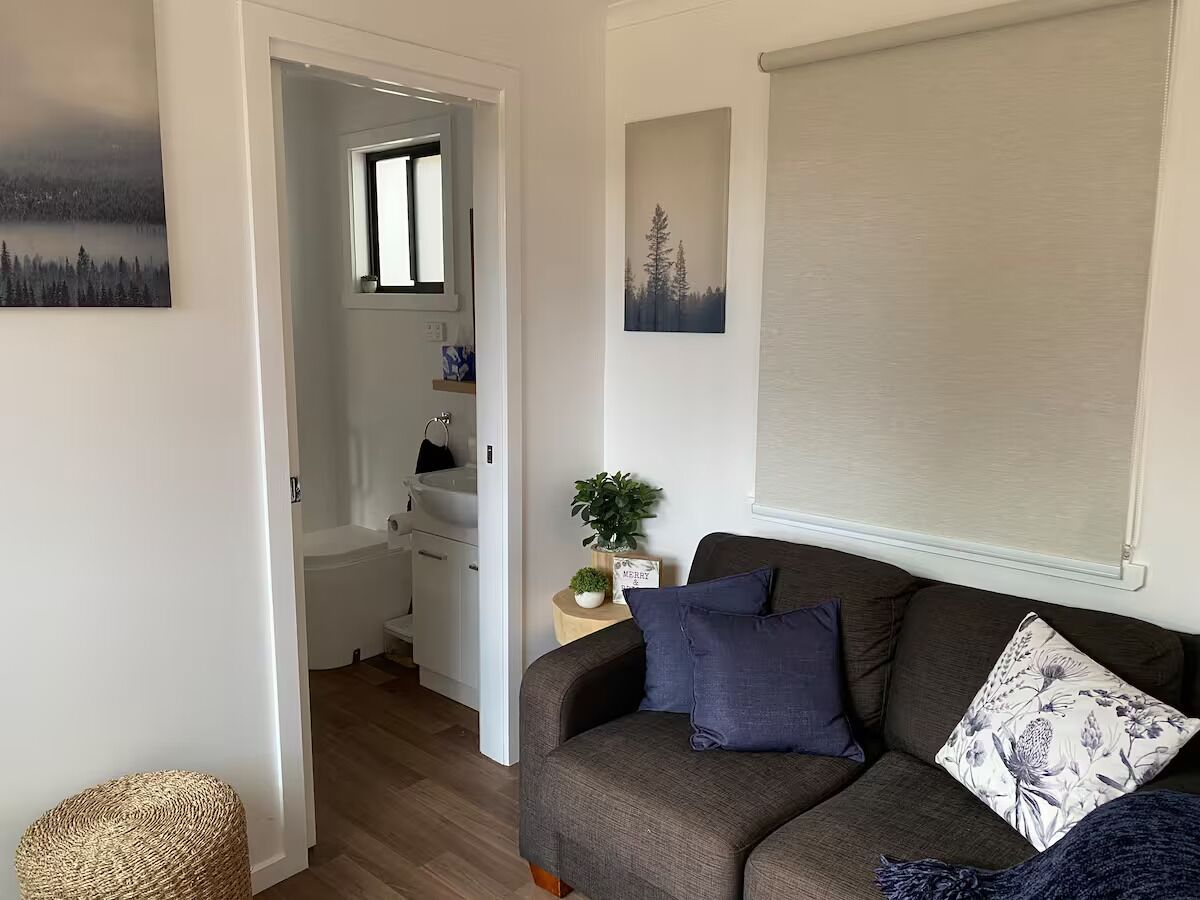 A cozy living room with a sofa, a small side table with a plant, and a window with blinds. A bathroom is visible in the background with a sink and a towel hanging.