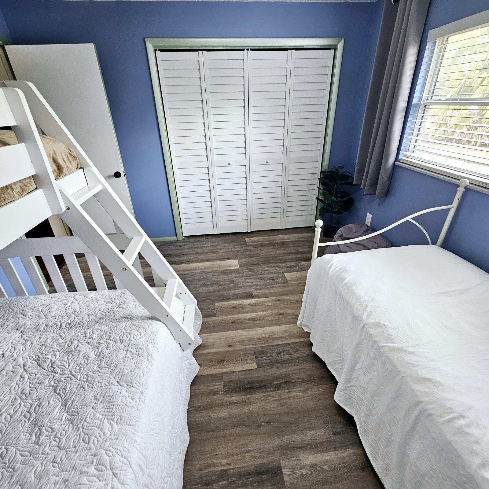 A bedroom with a wooden floor, two beds with white bedding, a white staircase, and a closet with white shutters.