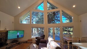 A spacious living room with a large brown leather sofa, a wooden coffee table, and a flat-screen TV. The room features a high ceiling with skylights and a view of trees outside.