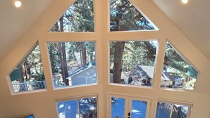 A spacious loft with a high ceiling, large windows offering views of a wooded area, a cozy seating area with a sofa and chairs, and a dining table.