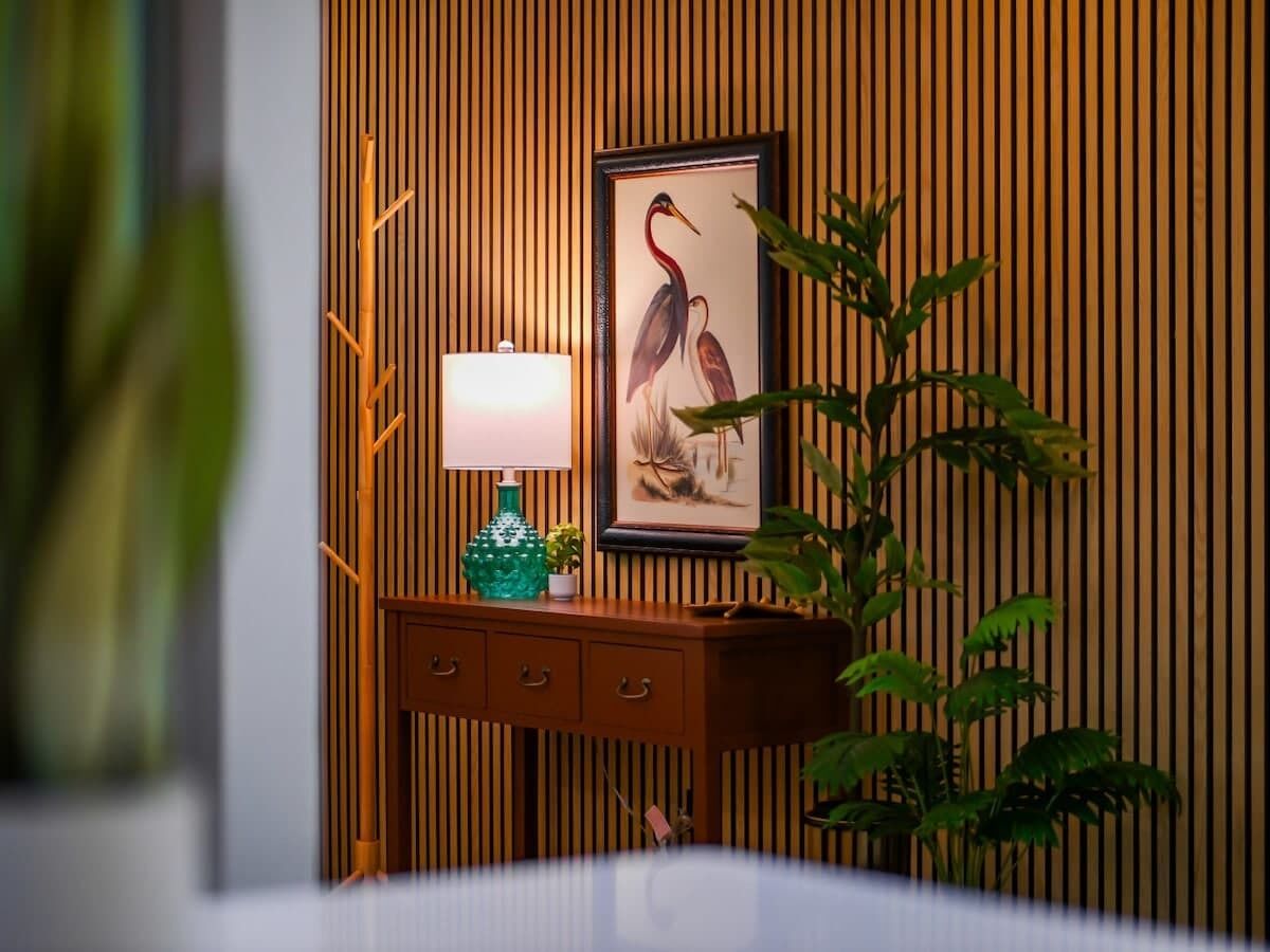 A room with a wooden slat wall, a framed artwork of a bird, a teal vase, and a bedside table with drawers.