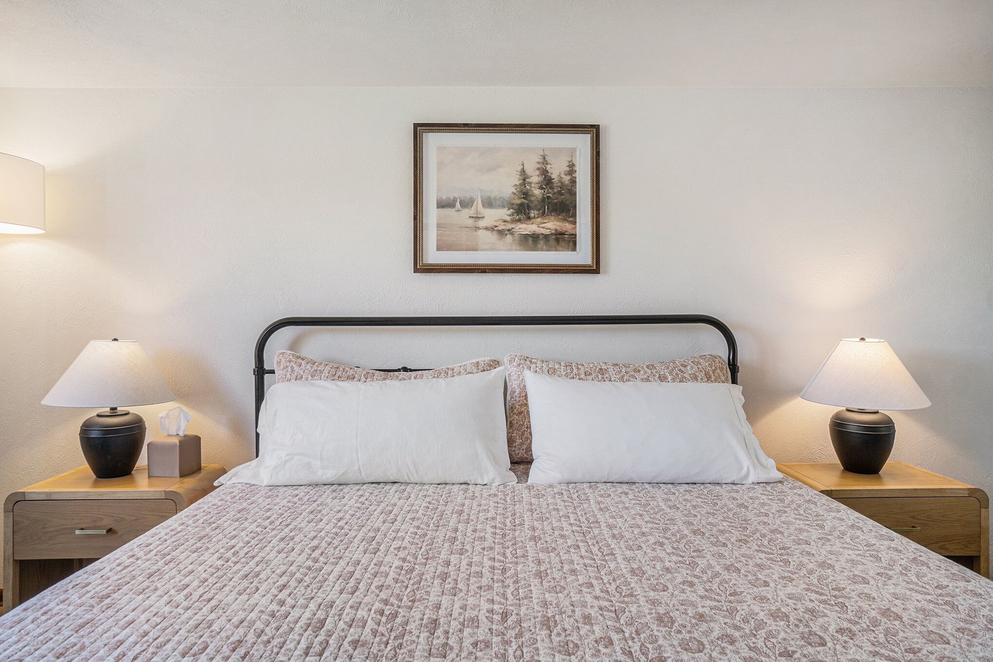 A neatly made bed with a patterned bedspread, two bedside lamps, a framed picture on the wall, and a small bedside table with a tissue box.
