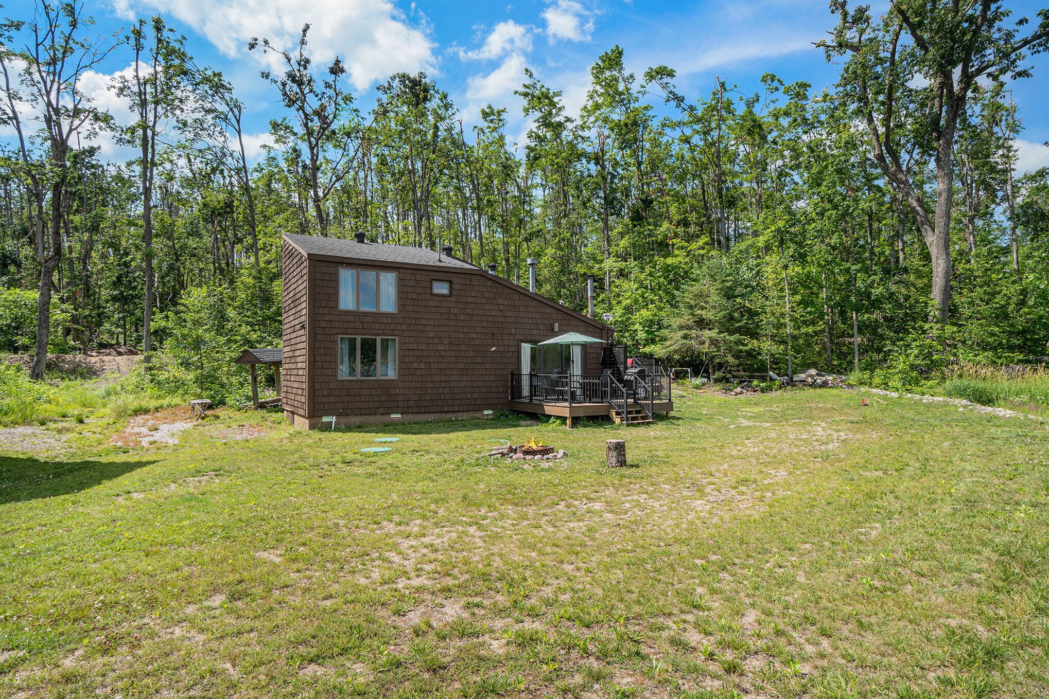 A single-story wooden cabin with a deck, surrounded by a grassy yard and dense forest.