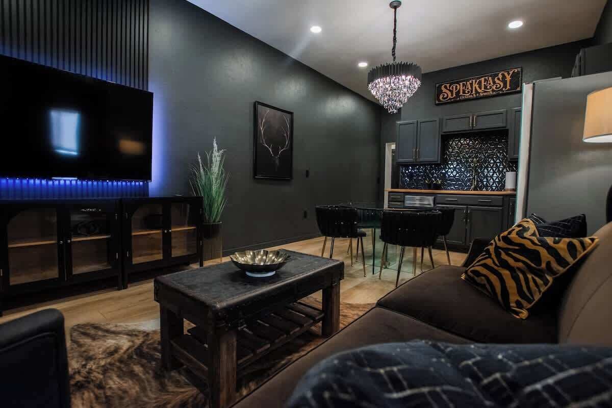 A modern living room with a flat-screen TV, a dining area with a chandelier, and a kitchen with a sign that reads "SPEAKASY."