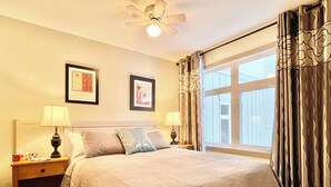 Un lit bien fait, recouvert d’une couvre-lit à motifs, deux lampes de chevet, un ventilateur de plafond et une fenêtre avec des rideaux.