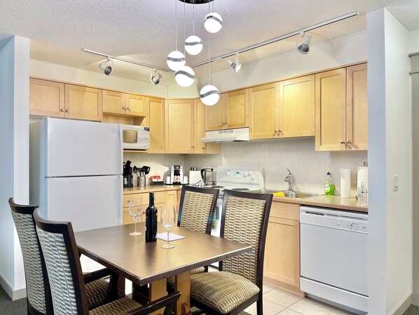 A modern kitchen with wooden cabinets, a white refrigerator, a microwave, a stove, and a sink. There is a dining table with chairs and a chandelier above.