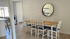 A spacious dining area with a wooden table and white chairs, a round mirror on the wall, and a view of the kitchen through an open door. - Farm cottage in 2.3 acres. Fully renovated for your comfort.  (Newmerella)