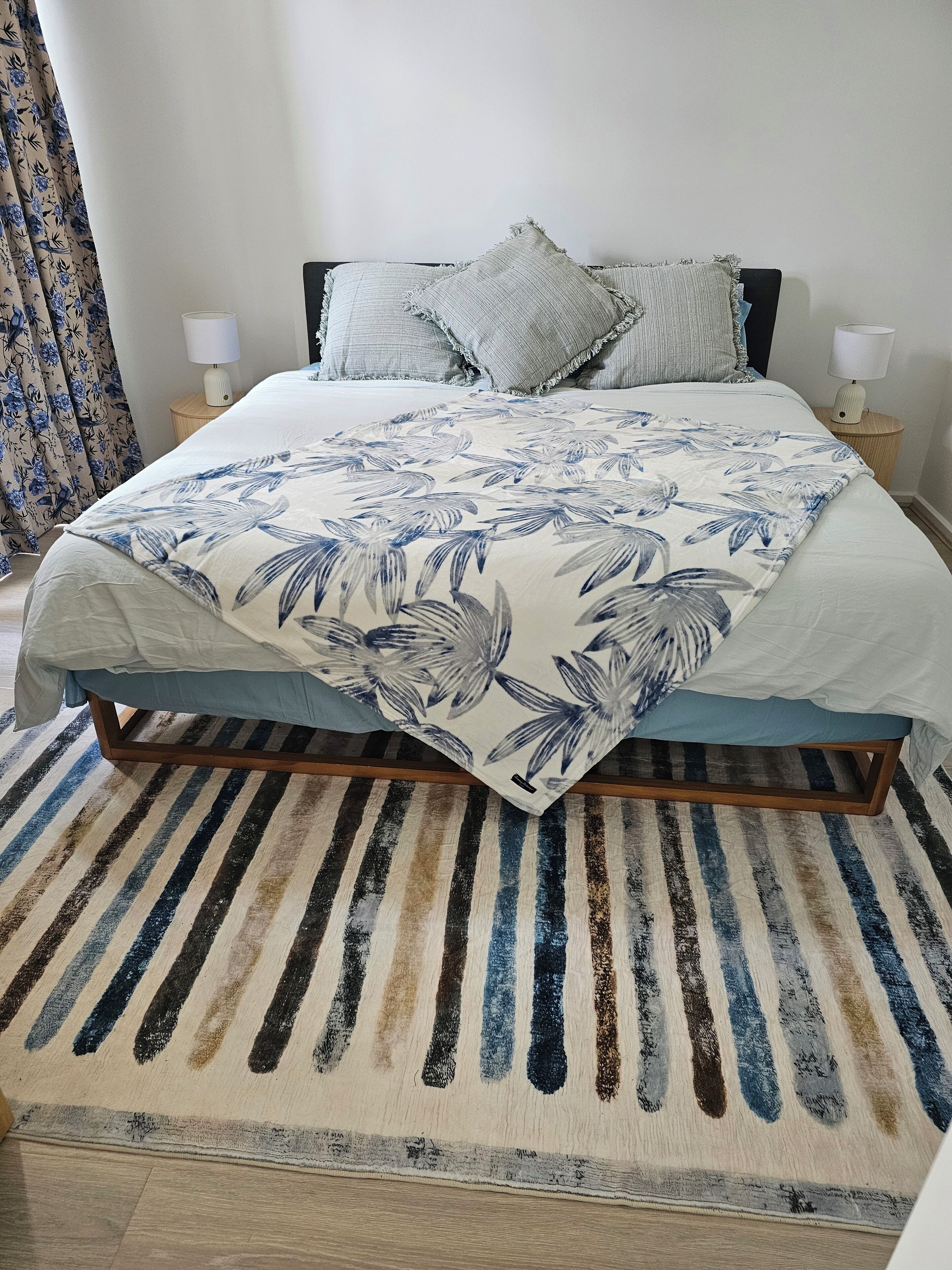 Una cama con una colcha estampada, dos almohadas, un tapete a rayas y una mesita de noche con una lámpara.