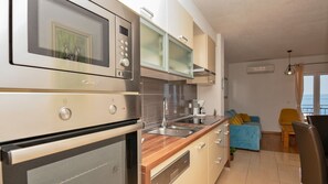 A modern kitchen with stainless steel appliances, a wooden countertop, and a built-in oven. There is a dining area with a blue sofa and yellow chairs.
