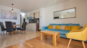 Una sala moderna con un sofá azul, un sillón amarillo y una mesa de centro de madera. En el fondo, se ve un comedor con sillas y una mesa.