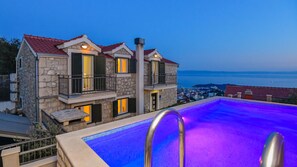 A two-story stone building with a swimming pool, balconies, and a view of the sea.