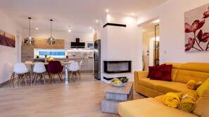 Una sala de estar moderna con un sofá amarillo, una zona de comedor con sillas blancas, una cocina con horno empotrado y una chimenea.