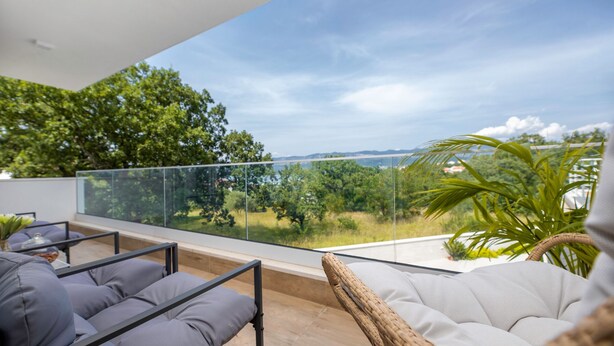 A balcony with a glass railing, wicker furniture, and a view of trees and hills.