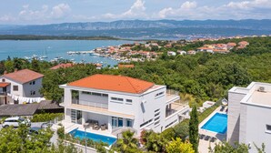 A modern two-story building with a swimming pool, surrounded by greenery and a view of a marina and distant mountains.