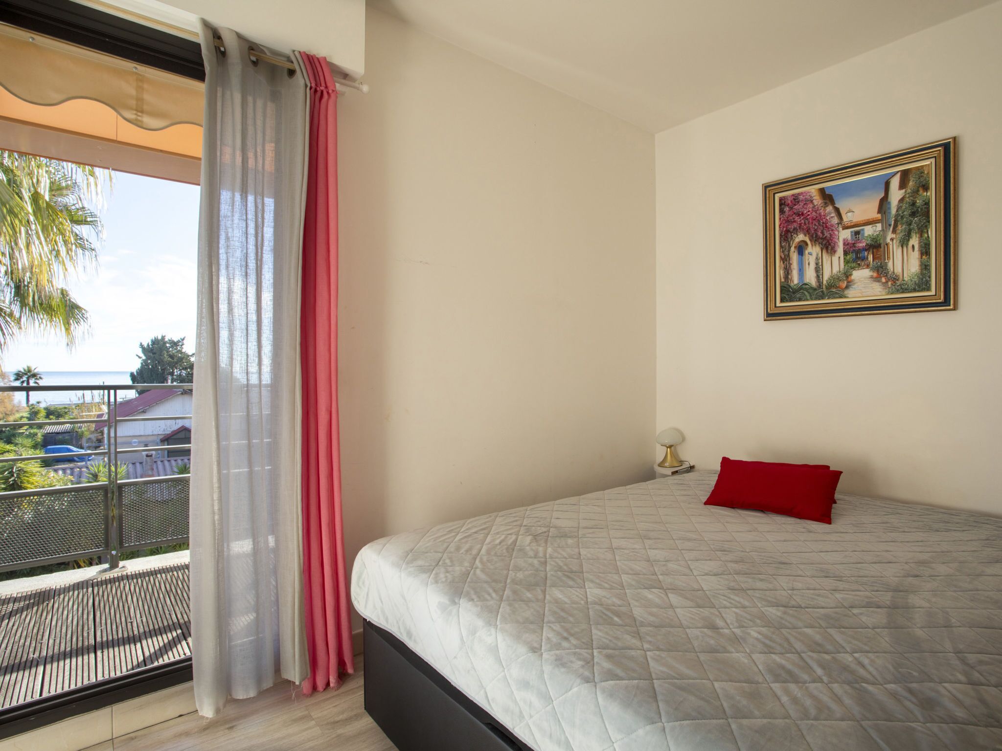 A bedroom with a bed, a painting, and a balcony with a view of the sea.