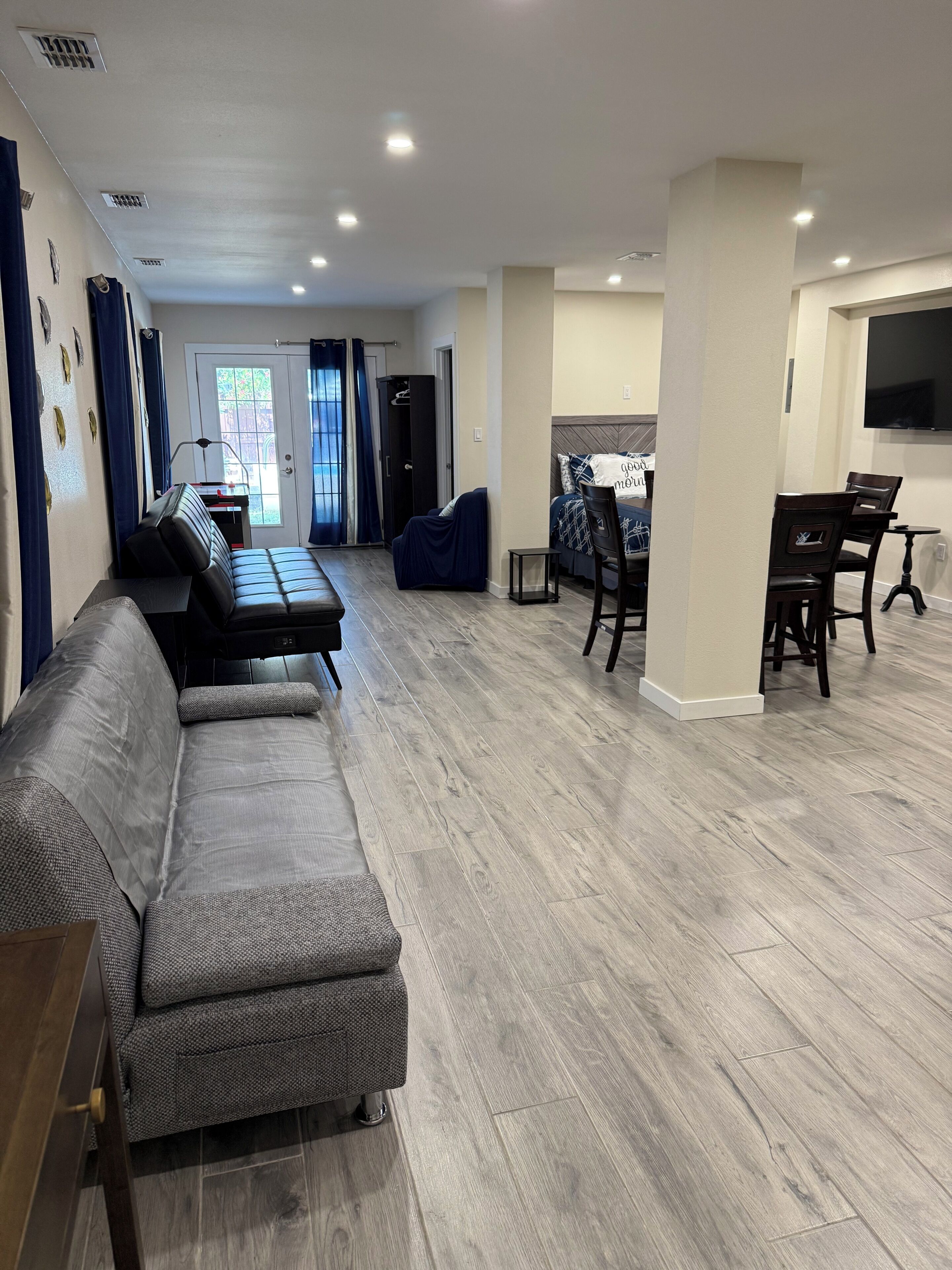 A spacious living area with a sofa, a dining table with chairs, a TV mounted on the wall, and a door leading outside.