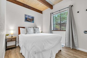 Une chambre à coucher avec un lit, une table de chevet, une lampe, une fenêtre avec des rideaux et un tableau au mur.