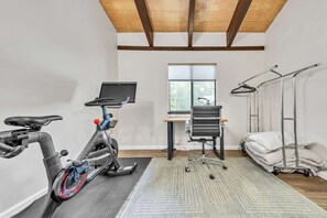Une pièce comprenant un vélo d’appartement, un bureau avec un ordinateur, une fenêtre avec des stores et un lit avec des oreillers.